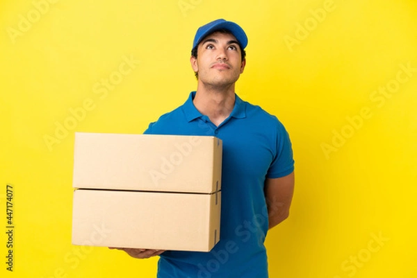 Fototapeta Delivery man over isolated yellow wall and looking up