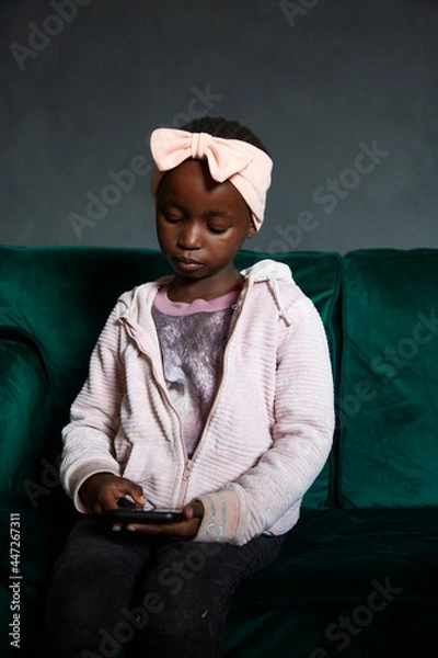 Fototapeta Low-key portrait of beautiful black South African girl wearing pink outfit with a pink bow on her head on a dark charcoal background, using phone while sitting on emerald green couch.