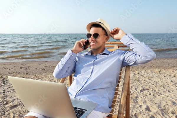 Fototapeta Happy man with laptop talking by mobile phone on beach. Business trip