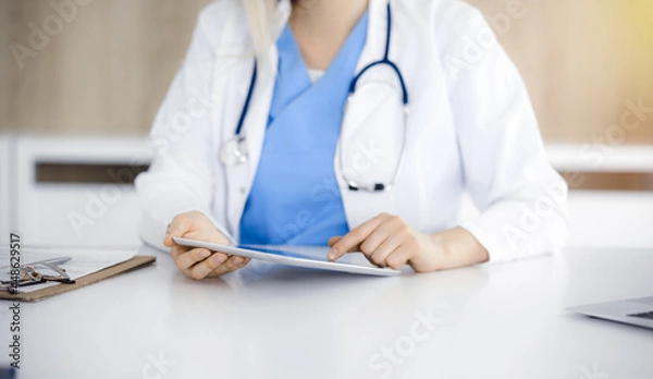 Fototapeta Unknown woman-doctor sitting in sunny clinic and using tablet pc, close-up. Data and best service in medicine