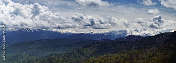 Fototapeta panoramic view of the snow-capped mountains at the foot of the forest against the backdrop of bright blue clouds