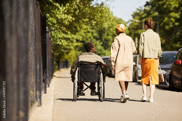 Fototapeta Rear view of handicapped black man in wheelchair walking with able-bodied friends and ommunicating with them