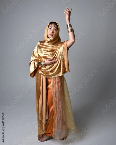 Fototapeta Full length portrait of pretty young asian woman wearing golden Arabian robes like a genie, standing pose holding flowing fabric, isolated on studio background.