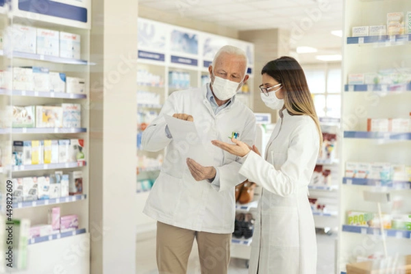 Fototapeta Education in pharmacy and corona virus. A mature pharmacist explains the documentation in the pharmacy to a young pharmacist in professional training. They wear uniforms and face masks