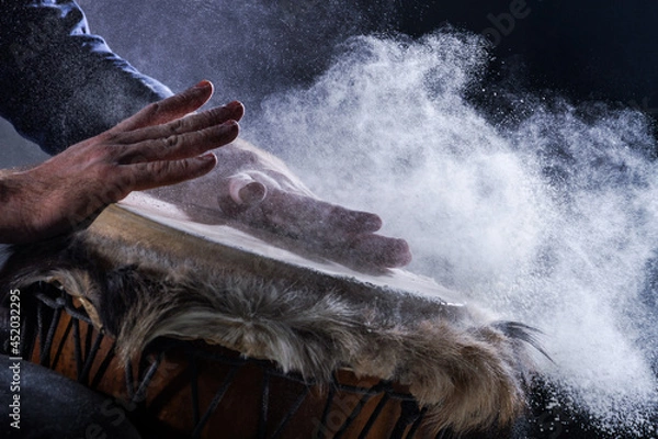 Fototapeta Man is playing on djembe drum, covered with talcum powder. Flour splashes on dark background. Summer festival concert performance. Ethnic rhythm. Percussion musical instruments and culture concept.