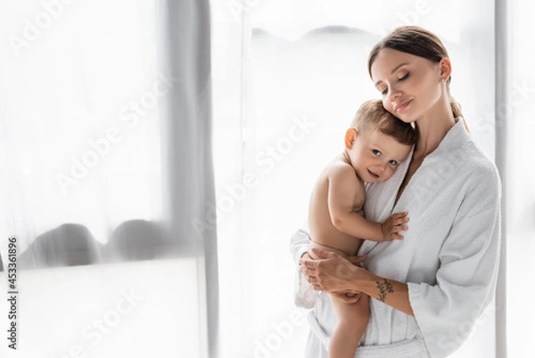 Fototapeta pleased and tattooed mother in bathrobe holding in arms nude toddler son