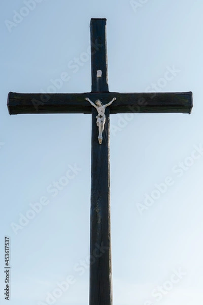 Fototapeta An old wooden Christian cross against a clear blue sky on a beautiful summer day. A statuette of Jesus in the center of the cross. The crucifixion of Christ. The feast of Easter and Resurrection.