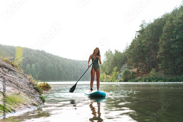 Fototapeta The slim young woman in green sweemsuit on sup boat with oar floating on river, weekend trip and local travel