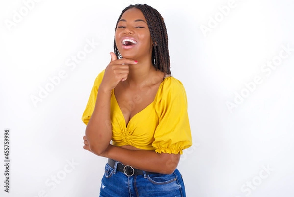 Fototapeta Young african american  woman with braids over white wall  laughs happily keeps hand on chin expresses positive emotions smiles broadly has carefree expression