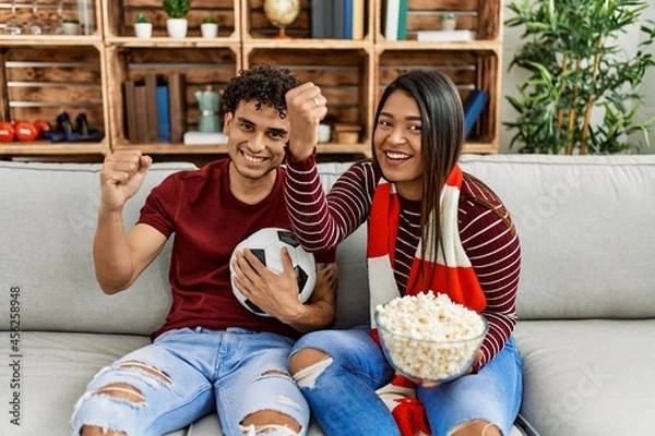 Fototapeta Young latin couple watching and supporting football match sitting on the sofa at home.