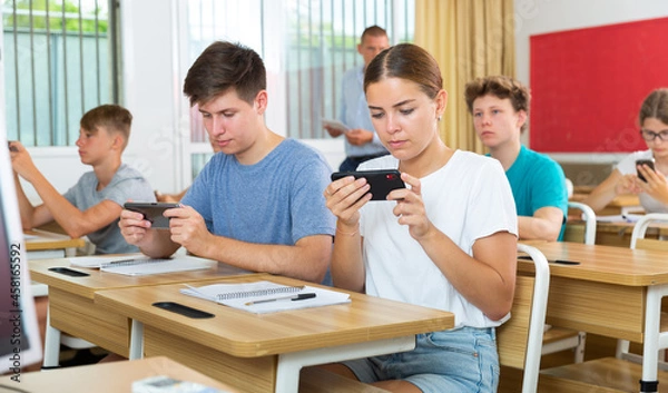 Fototapeta Young students with smartphones sitting in class room. Teacher explaining something to them.