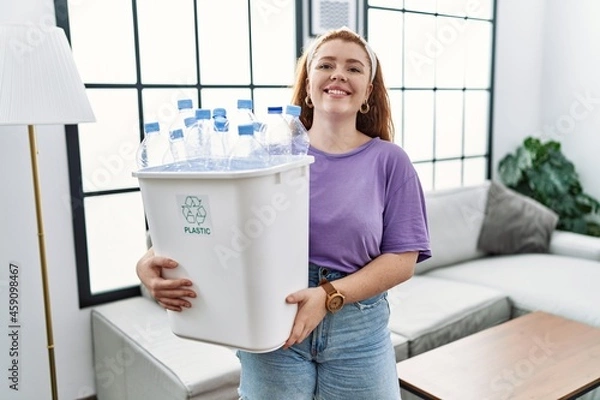 Fototapeta Young redhead woman holding recycling wastebasket with plastic bottles with a happy and cool smile on face. lucky person.