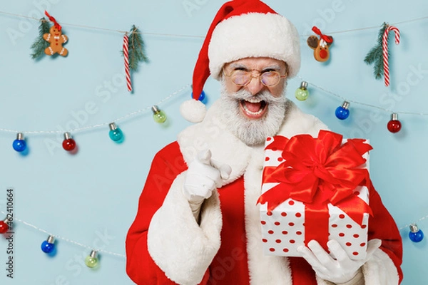 Fototapeta Old Santa Claus man in Christmas hat red suit hold present box gift ribbon bow point finger camera on you scream isolated on plain blue background studio. Happy New Year 2022 merry ho x-mas concept.