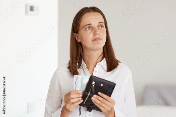 Fototapeta Indoor shot of sad upset dark haired female wearing white casual style shirt, holding wallet with money in hands and looking away, expressing sadness, have not enough money for shopping.