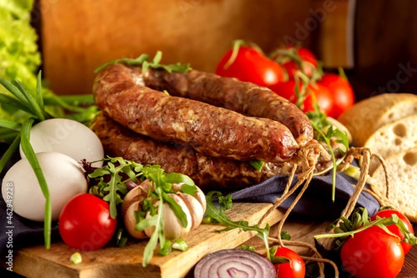 Fototapeta Homemade sausage - tasty traditional celebration food on old wooden table with vegetables: onions, garlic, cucumbers, tomatoes, arugula, eggs.