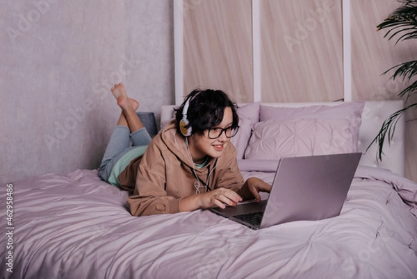 Fototapeta a Caucasian teenage girl is lying on the bed with headphones and a laptop