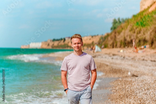 Fototapeta A young blond man walks on the beach near the azure ocean or sea. The concept of a happy summer vacation