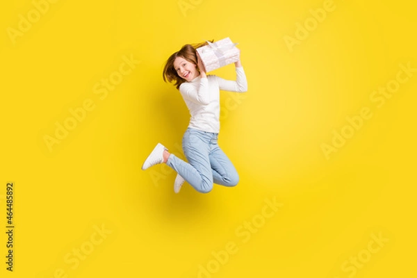 Fototapeta Full length photo of happy nice young small girl jump up hold gift box smile isolated on yellow color background