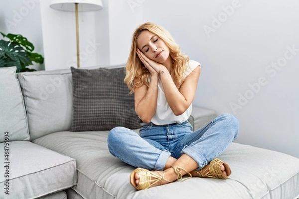 Fototapeta Beautiful blonde woman sitting on the sofa at home sleeping tired dreaming and posing with hands together while smiling with closed eyes.