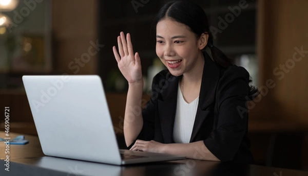 Fototapeta Image of happy asian woman wearing white shirt smiling and waving hand at laptop, while speaking or chatting on video call in office.