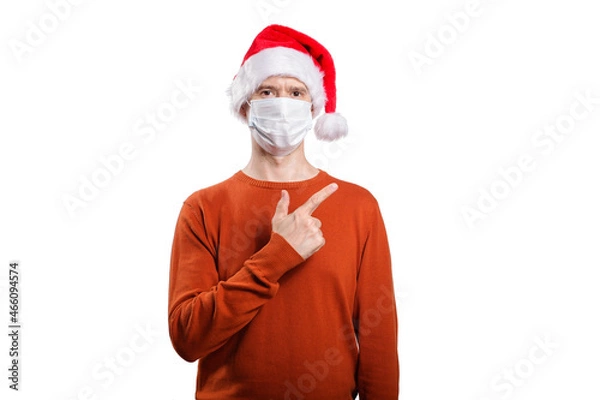 Fototapeta an isolated shot of a happy man in a medical mask from the coronavirus, in casual clothes, an orange sweater, a Christmas hat. With his finger he points to the right, looks into the camera