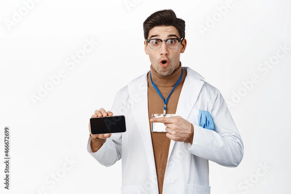 Fototapeta Image of surprised doctor pointing at mobile phone screen horizontal, showing application on smartphone, standing over studio background