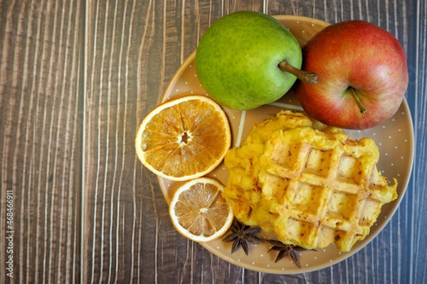 Fototapeta pumpkin Belgian gluten-free waffles lie on a brown plate with a dry slice of lemon, pumpkin on a background of yellow maple leaves on a wooden table with a pear and an apple. top view. gluten-free