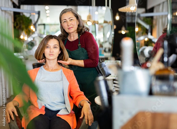 Fototapeta Skilled aged female hairdresser offering modern hairstyling to young woman client visiting professional hair salon.