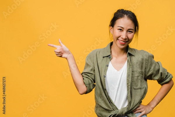 Obraz Portrait of young Asian lady smiling with cheerful expression, shows something amazing at blank space in casual cloth and looking at camera isolated over yellow background. Facial expression concept.