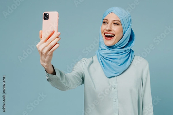 Fototapeta Young smiling fun arabian asian muslim woman in abaya hijab doing selfie shot on mobile cell phone isolated on plain blue background studio portrait People uae middle eastern islam religious concept