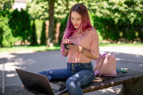 Fototapeta Woman writing in a notebook sitting on a wooden bench in the park. Girl working outdoors on portable computer, copy space.