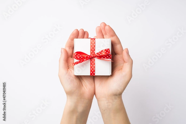 Fototapeta First person top view photo of young woman's hands holding small white giftbox with red dotted ribbon bow in palms on isolated white background