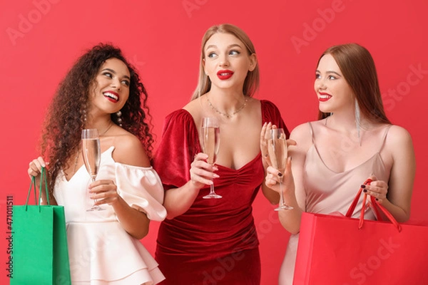 Fototapeta Beautiful young women with glasses of champagne and shopping bags on color background