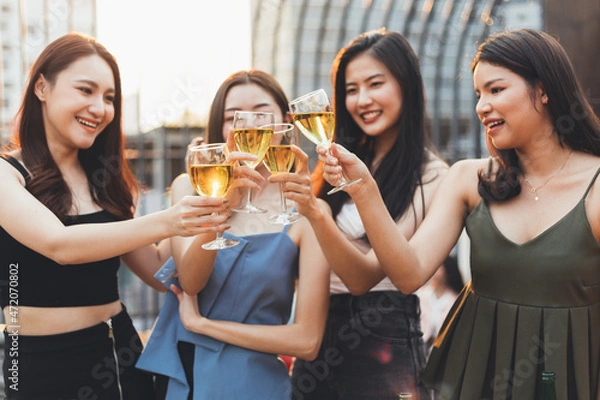 Fototapeta Group of happy smiling Asian girl friends celebrating party with champagne toasting drinks at rooftop cafe together, female gang chatting, laughing, night lifestyle of young people, focus on glasses