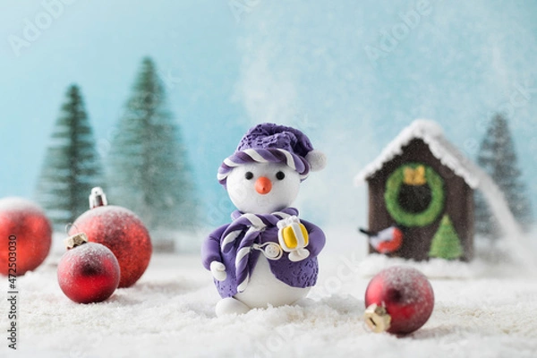 Fototapeta Toy snowman and red balls on fake snow near small house in decorative forest