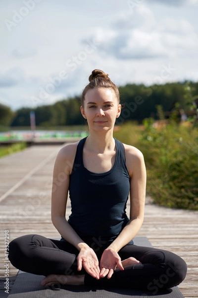 Fototapeta Young relaxing smiling woman practicing yoga on the wooden pier near lake.