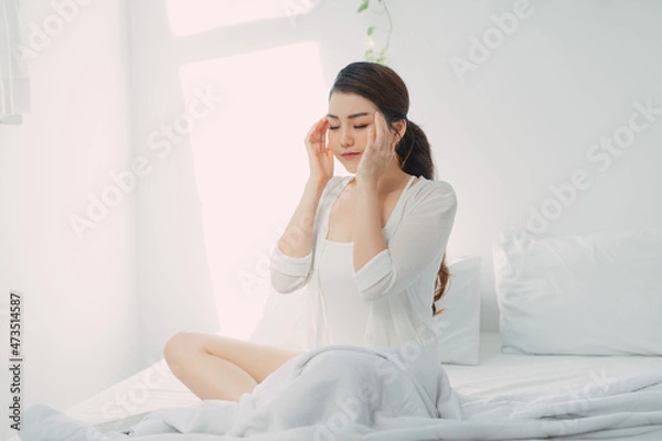 Fototapeta Young woman with headache, she is sitting on the bed and touching her temples