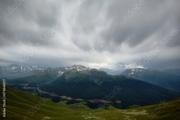 Fototapeta snow-capped mountain peaks, a road under construction in a green mountain valley. Gray cloudy day