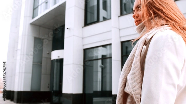 Fototapeta A girl in a white coat and a scarf is walking down the street, smiling. Business center in the background. Success