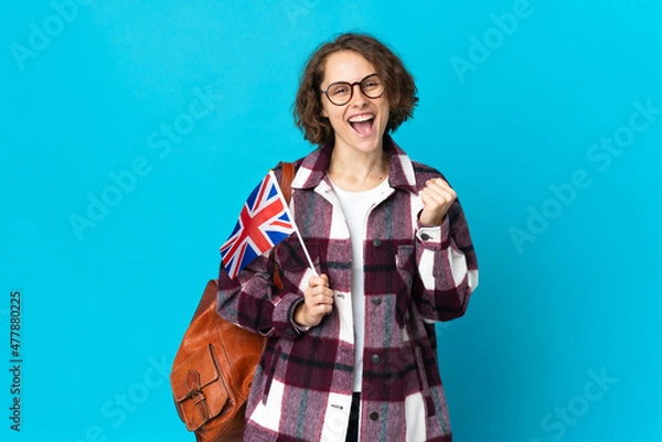 Fototapeta Young English woman holding an United Kingdom flag isolated on blue background celebrating a victory in winner position