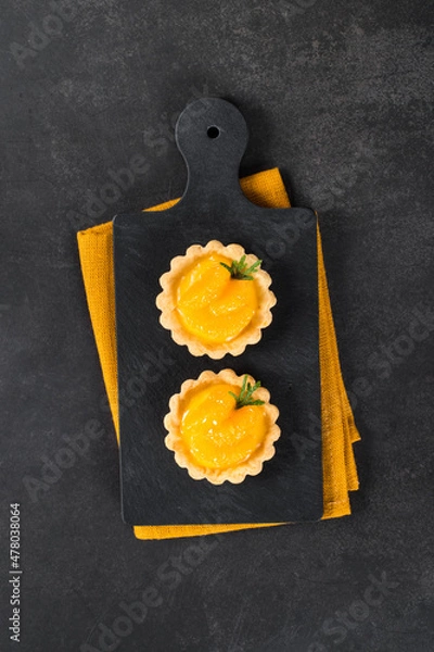 Fototapeta Twoo Tartlets with custard and slices of tangerines covered with syrup on a serving board on a linen napkin on a dark gray background. Top view