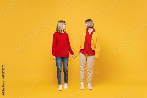 Fototapeta Full body smiling friendly happy cool woman 50s in red shirt have fun with teenager girl 12-13 years old. Grandmother granddaughter look to each other hold hands isolated on plain yellow background