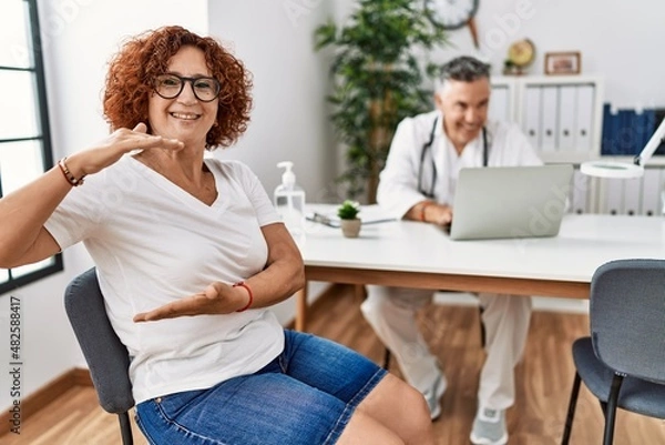 Fototapeta Senior woman sitting at doctor appointment gesturing with hands showing big and large size sign, measure symbol. smiling looking at the camera. measuring concept.