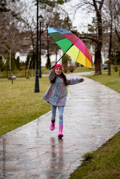 Fototapeta A cute little girl in a blue cape, pink boots and a pink hat runs through puddles and has a fun.The girl has a rainbow umbrella in her hands. Happy childhood. Early spring. Emotions.