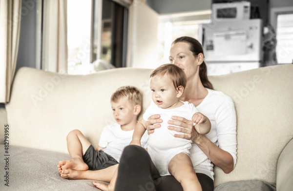 Fototapeta Mother and her children at home sitting on couch sofa watching TV, videos 