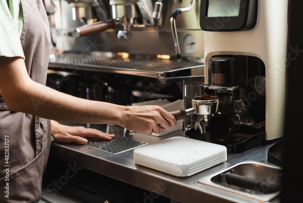 Fototapeta Cropped view of barista in apron holding portafilter near coffee grinder in cafe.