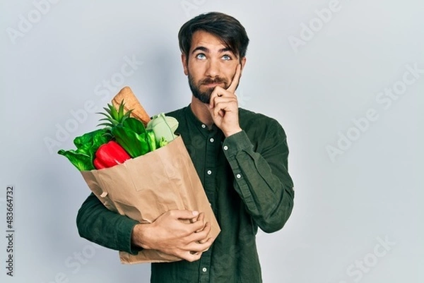 Fototapeta Young hispanic man holding paper bag with bread and groceries serious face thinking about question with hand on chin, thoughtful about confusing idea