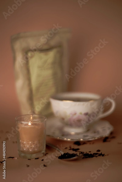 Fototapeta a cup of white and pink with flowers with black tea with tea packaging and loose tea and a burning pink candle in a drain with pearls on a pink background