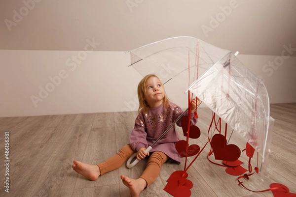 Fototapeta a blonde girl is holding an umbrella in her hands, sitting on the floor and looking at the hearts that hang from the umbrella