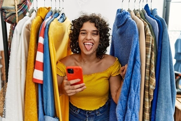 Fototapeta Young hispanic woman searching clothes on clothing rack using smartphone sticking tongue out happy with funny expression. emotion concept.